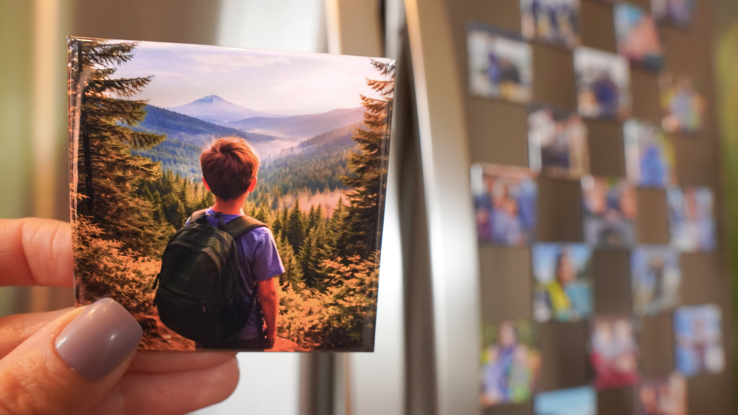 Hand holding a custom 2.5 inch square photo magnet of a person in a mountain landscape, with a fridge full of photo magnets in the background