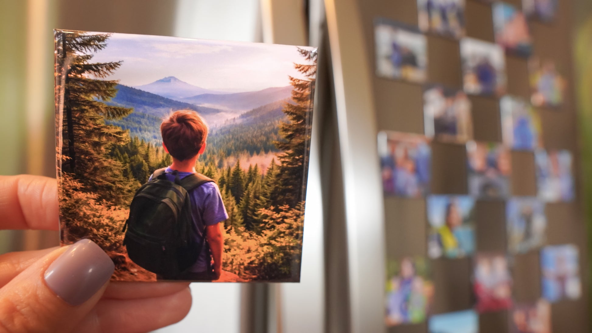 Hand holding a custom 2.5 inch square photo magnet of a person in a mountain landscape, with a fridge full of photo magnets in the background